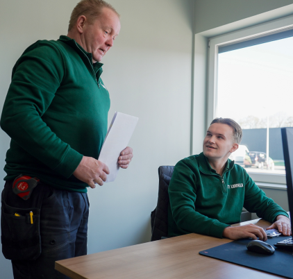 Twee mannen zitten op kantoor. De een staat met papieren, draagt een groene trui en gereedschapsriem, die doet denken aan de veerkracht van een lamel. De ander zit aan een bureau met een toetsenbord en kijkt omhoog naar de staande man. Beiden dragen vergelijkbare groene tops. Een raam aan de rechterkant toont een heldere dag buiten.