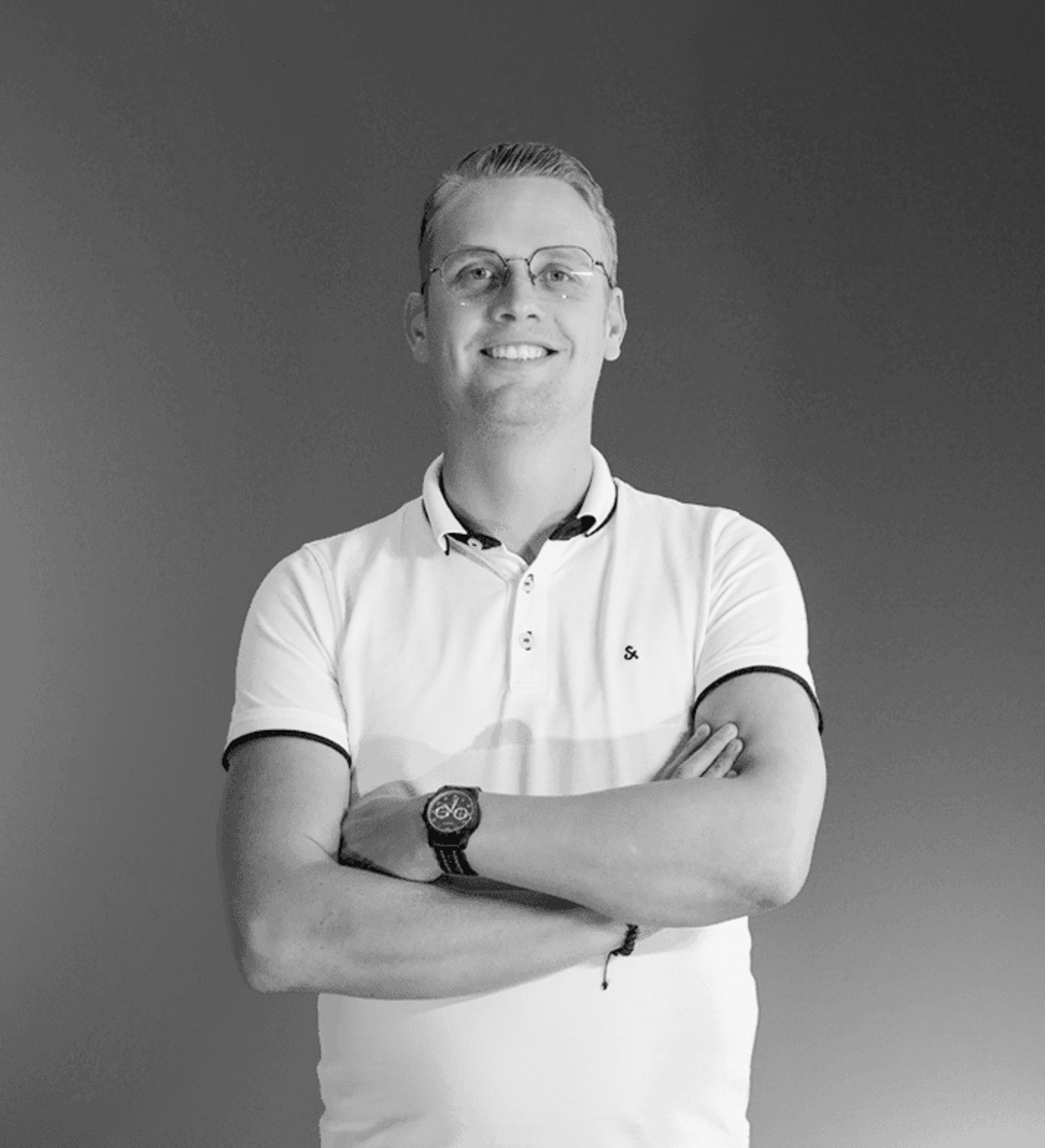 Over ons 2 Een lachende man met kort haar en bril staat tegen een effen achtergrond. Hij draagt een witte polo met donkere bies en een polshorloge en slaat zijn armen nonchalant over elkaar, wat de laagdrempelige stijl van rijnders webdesign uitstraalt. De foto is zwart-wit.