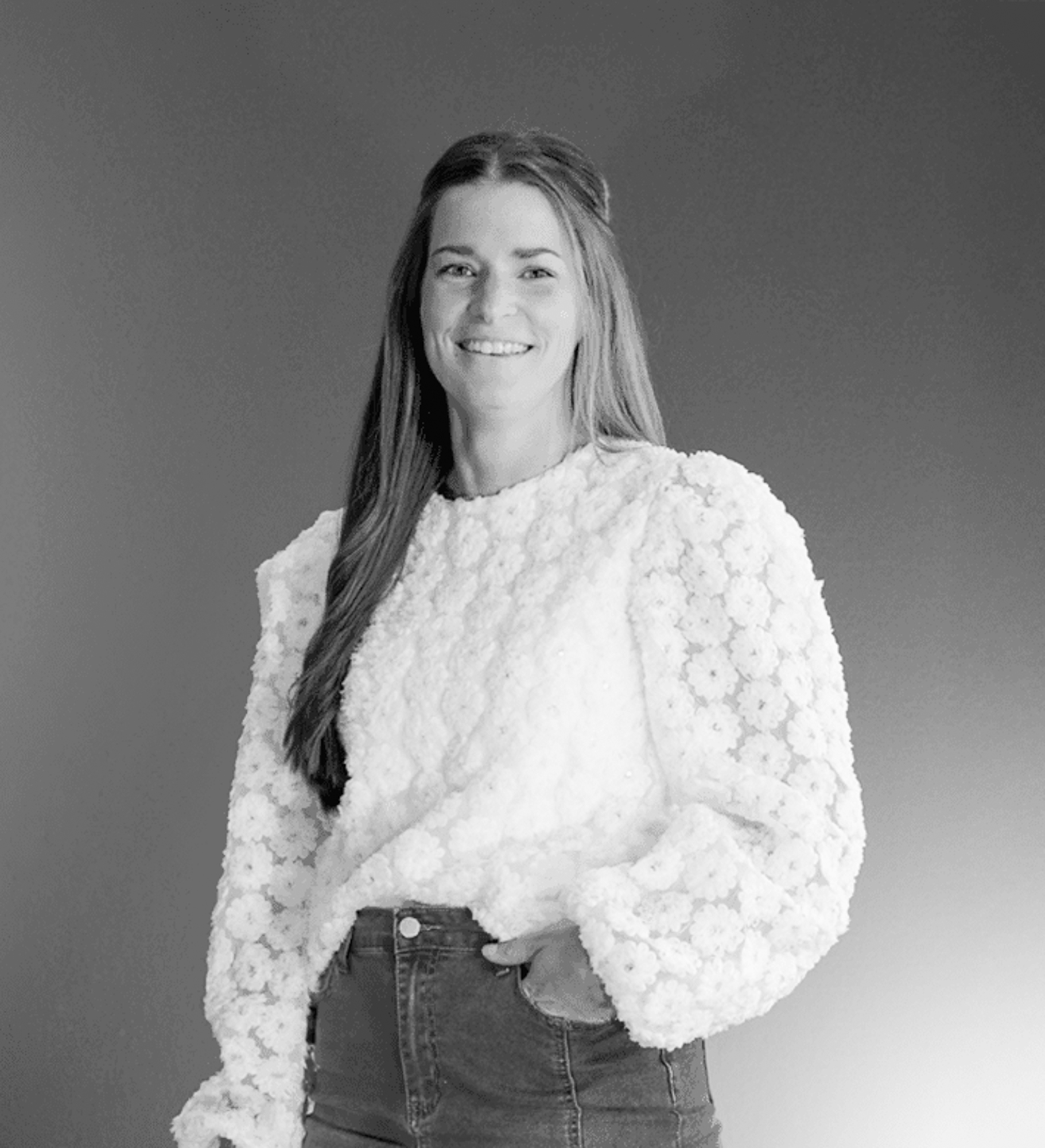 Over ons 4 Een vrouw met lang steil haar lacht naar de camera en draagt een blouse met bloemmotief en een broek met hoge taille. Haar stijl weerspiegelt de moderne elegantie van rijnders webdesign tegen een effen, zwart-witte achtergrond met kleurverloop.