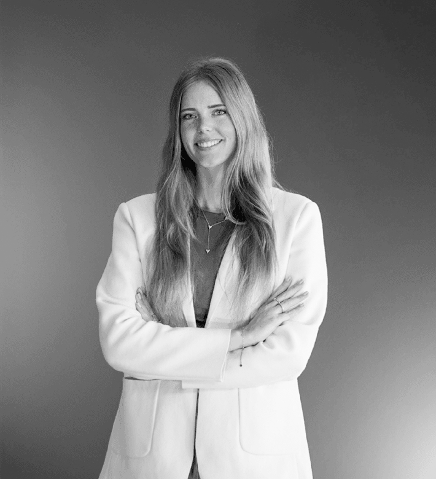 Over ons 6 Een vrouw met lang, steil haar staat tegen een effen achtergrond en glimlacht zelfverzekerd met haar armen over elkaar. Haar witte blazer en subtiele sieraden geven haar een moderne look - perfect voor de strakke esthetiek van rijnders webdesign. De afbeelding is in zwart-wit.