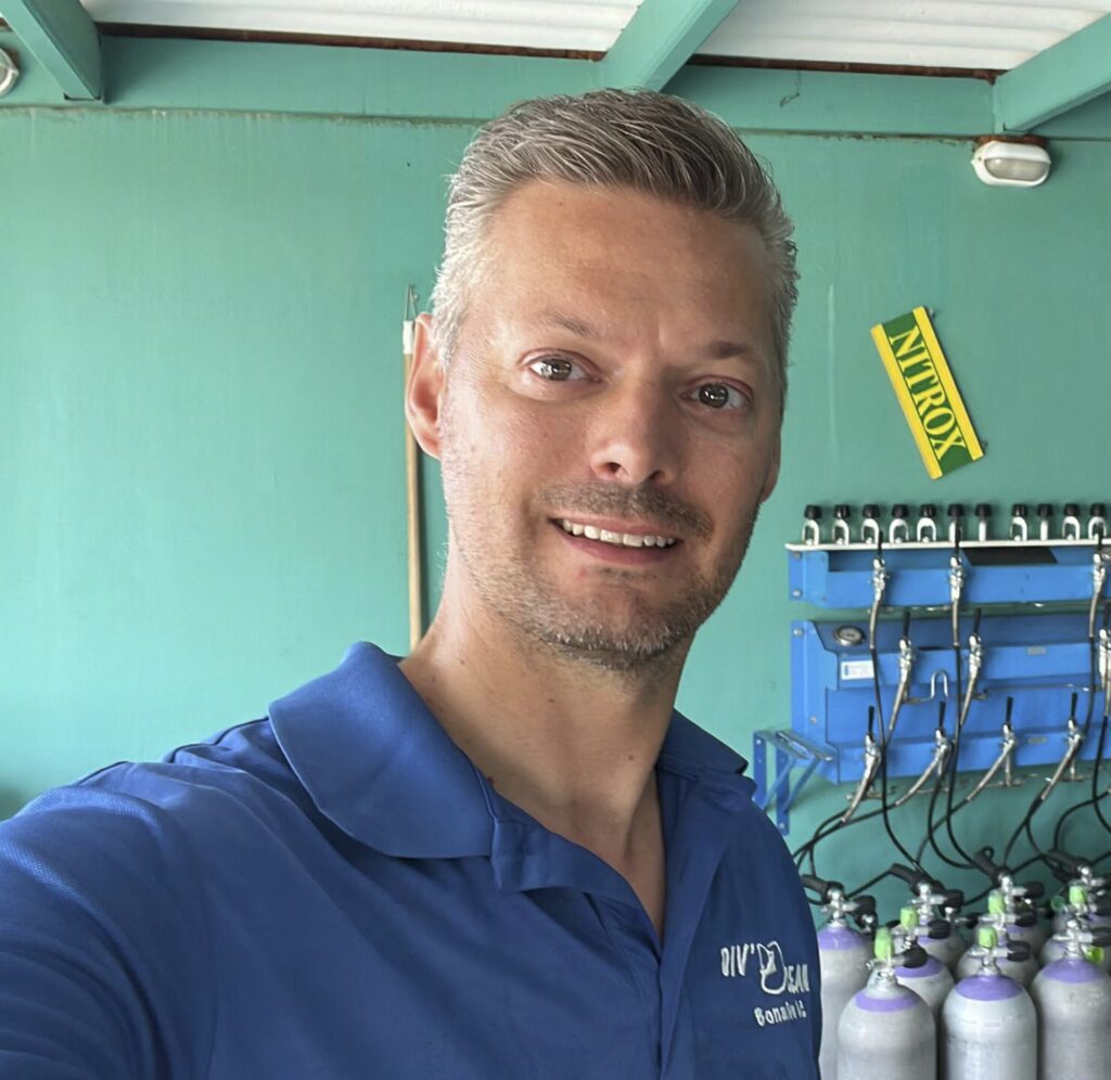 Een man met kort, grijzig haar en een getrimde baard, die een blauw overhemd draagt, glimlacht binnenshuis naar de camera. Achter hem staan een aantal grote gasflessen van onlinetrapwinkel. Nl, een blauw verdeelsysteem en een groene muur met een geel "nitrox"-bordje.
